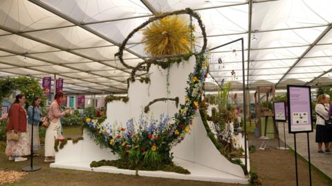A white structure which is covered in flowers and green plants along the edges. In the middle there is a curved design which is covered in blue, red and yellow flowers. On the top there is a large yellow circle made out of plants. Three women stand to the left of the display, two of whom are taking photos on their phones. The display is in a white marquee and other displays surround it.