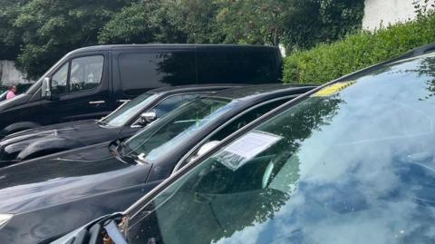A close-up of a car windshield with a notice attached to it, other black cars in the background also have notices on them.