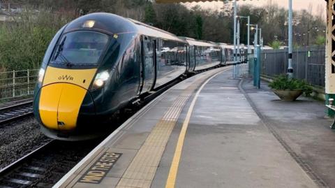 A green GWR train pulls into a railway station. It has its lights on. The platform is empty.