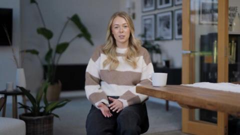 A blonde woman wearing a brown and white jumper with wide stripes and black trousers. She is sitting next to a table on top of which is a white mug