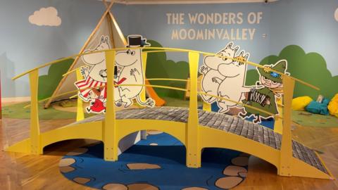 A brightly colored indoor play exhibit themed “The Wonders of Moominvalley” features a small yellow bridge with cartoon Moomin characters and a tent-like play area in the background.