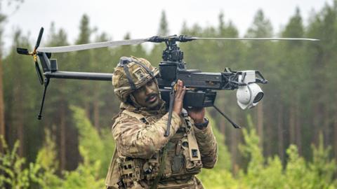 A British soldier in battle fatigues carries a a single-rotor helicopter style Ghost drone on his shoulder in a forest