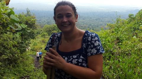 Jo Trevor at a green area of Congo. She is wearing a dark blue dress with white flowers on. Two other people can be seen walking down the path behind her. It is overcast.