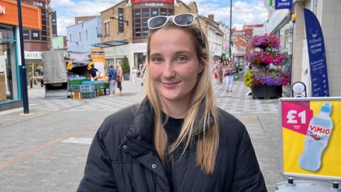 A blonde woman in a black puffer jacket and a pair of sunglasses resting on her head. She is standing at a high street.