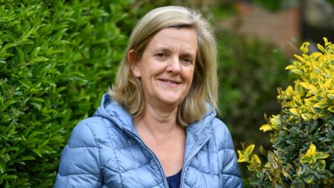 A blonde woman in a blue coat standing in between two bushes.