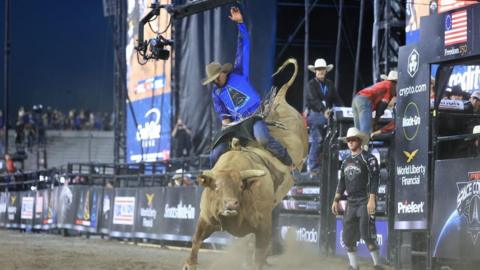 A professional bull rider, wearing a cowboy hat and blue shirt featuring the triangular logo of US Space Force, clings on while riding on the back of a massive, brown horned bull in a stadium filled with dirt 