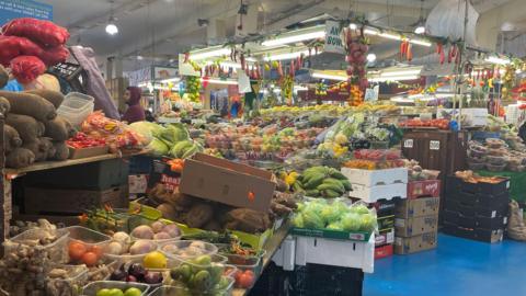 A large stall at an indoor market covered with fruit and vegetables with boxes of them piled up alongside. A man can be seen looking around on the left.
