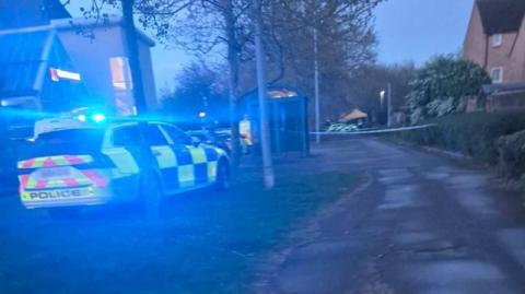 Police cars and vans parked on a street. There are several cars visible at either end of the street as it is starting to get dark. Police tape is also visible.