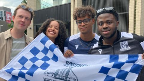 Niamh Bligh, who has curly brown hair, smiling at the camera. She is flanked by three men. They are all holding a blue and white chequered Southend flag.