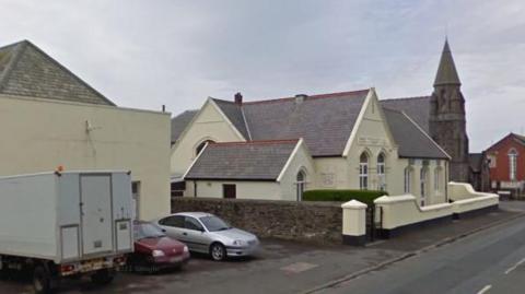 A set of cream coloured, peak roofed buildings on Station Road. There are cars parked outside on of them.