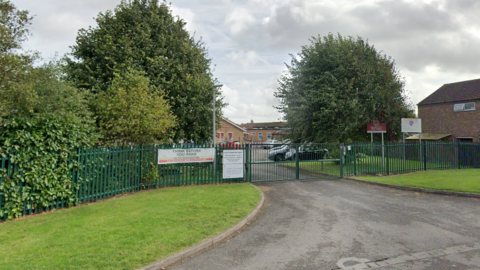 A school driveway flanked by green verges and a hedge. The school's sign is visible in the background. 