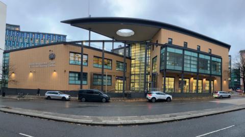 The front of Bristol Magistrates' Court, a building of three-storeys which is quite modern and built from a yellowy brick with blue-tinged windows. There are a few cars and a van waiting out the front of the building. It is a rainy day and the road looks wet.