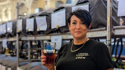 Anneli Baxter, general manager of the Loose Canon brewery in Abingdon  and regional chair of SIBA, holding a pint of beer. She is wearing a black T-shirt with her brewery's branding. There are barrels stacked in rows behind her.