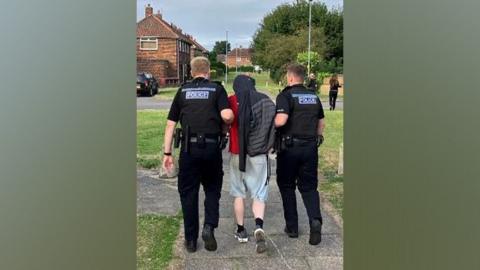 Two police officers dressed in black holding a man by his arms and leading him down a residential street. He's dressed in shorts and a red t-shirt and covering his head with a tracksuit top.