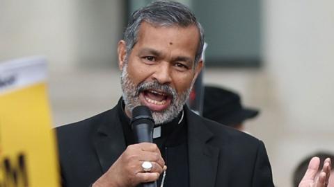 Right Reverend Dr John Perumbalath speaks into a microphone as he stands in a street