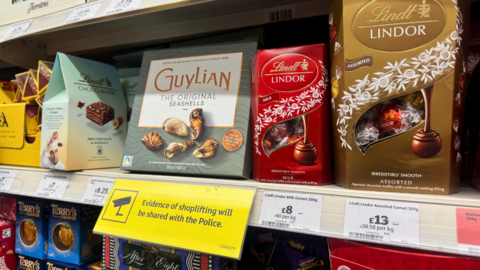 A supermarket shelf stocked with boxed chocolates from brands like Lindt, Guylian and Thorntons, with a yellow sign in the centre warning that evidence of shoplifting will be shared with the police.