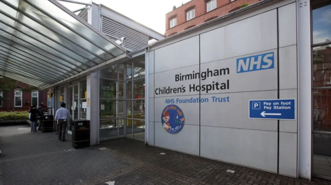 The hospital trust sign is visible by the entrance to the hospital with glass roofing and frontage.