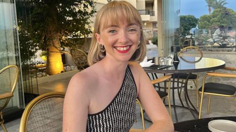 Billie Nicholls in a black and white top sitting at table on holiday. She has blonde shoulder-length hair and a fringe. She wears red lipstick.