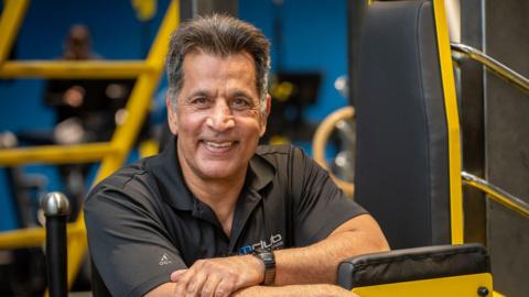 A man wearing a black polo shirt, is leaning on a yellow piece of gym equipment. He has short dark hair and smiles showing his teeth while his left hand rests on his right forearm.