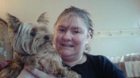Ann Green is cuddling a pet dog and sitting on a sofa. She is wearing a black top and has her hair tied back in a ponytail and is wearing earrings.