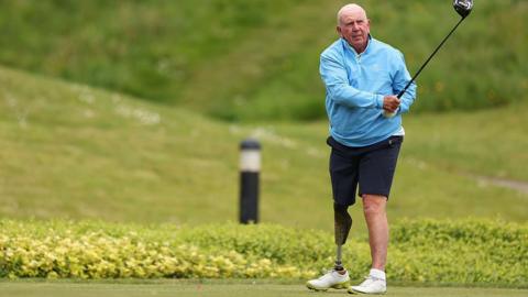 Richie Willis, dyn gwallt gwyn mewn top glas a shorts du, yn chwarae golff - coes brosthetig yw ei goes dde