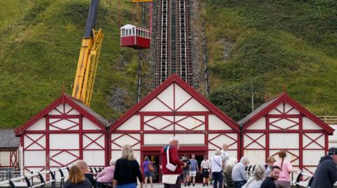 A crane is lowering a red and white carriage onto the tracks which descend down the grassy cliff. There are three red and white buildings at the bottom of the cliff, with people dotted around out front.