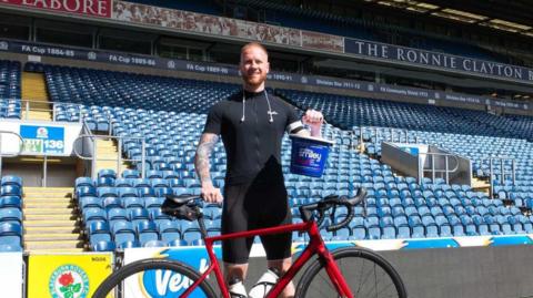 Lewis Robinson stands with his bike in Blackburn Rovers' Ewood Park stadium holding a charity collection bucket