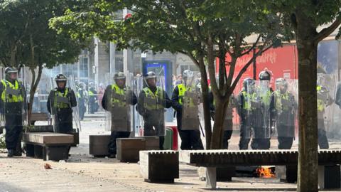 Police wearing protective equipment and carrying shields form a barrier in Hull city centre during riots on 3 August. Debris lies on the pavement with some of it on fire.