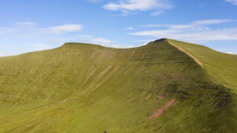 The green hills and slopes of Pen y Fan seen on a nice day. The sky is blue above the hills.