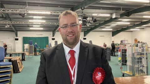 Matt Bishop, pictured in a hall where votes have been counted. He is wearing a suit with a red tie and a red rosette. 