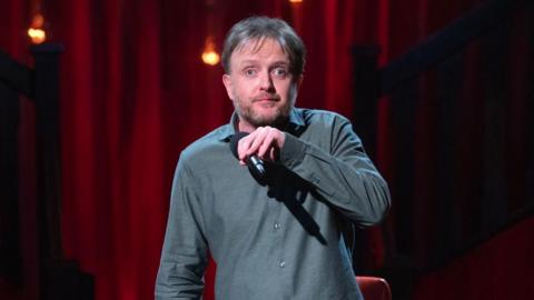 Comedian Chris McCausland stands on stage with a red curtain behind him. He is holding up a microphone. 