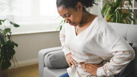 Stock photo shows a person looking down as they hold their lower stomach while sitting on a sofa at home.