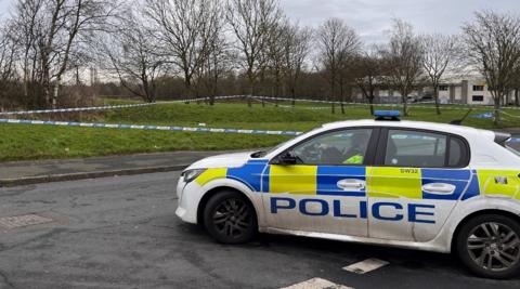 Police tape around a section of grass in the background. A police car is at a junction in the foreground.