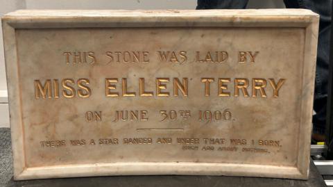 A beige marble stone in rectangle shape. The stone has gold text written on the front which reads "The stone was laid by Miss Ellen Terry on June 30th 1906" alongside a quote along the bottom.