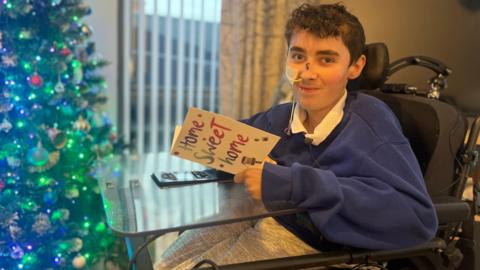 Curtis Herron is pictured sitting in his wheelchair next to the Christmas tree as he holds a "Home Sweet Home" card. He has dark, short curly hair and is wearing a blue jumper with a white shirt.