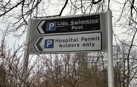 The image shows two parking signposts set against bare tree branches. The top sign is black with a blue parking “P” and the words “Lido Swimming Pool.” Beneath it, a second sign displays a blue “P” with the text “Hospital Permit Holders Only.”