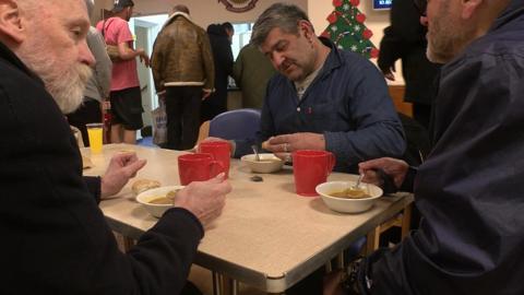 Three men sit at a table eating soup out of bowls. One is wearing a blue shirt, has an earring and grey hair. Another has a white beard and wears a black fleece. Another has a beard and a blue shirt. In the background people are queuing for food. 