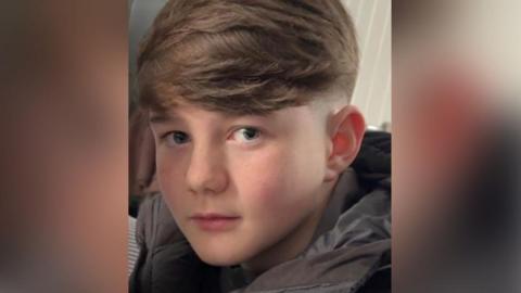 Twelve-year-old Junior Slater with short brown hair wearing a black coat. He is sitting on a sofa in a house.