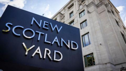 Exterior of the London Metropolitan Police building - the New Scotland Yard sign is visible in the near ground, with an imposing white stone five storey building behind it 