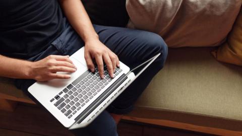 An unrecognisable man wearing a navy t-shirt and dark blue jeans is sitting on a sofa using a laptop.