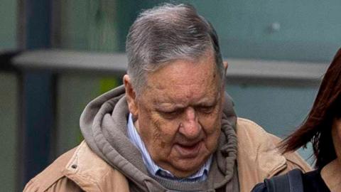 William Lloyd-Lavery entering court, it is a shot of him from the shoulders up. He has grey hair and is wearing a tan jacket over a grey hoodie and blue and white striped shirt. He is looking down towards the ground.
