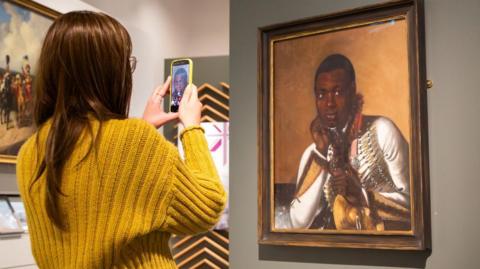 A woman wearing a yellow jumper takes a photo of a painting. The work depicts a black military bandsman holding a cymbal.