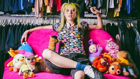 Emma Rogue sat on a large pink chair with cuddly toys beside her on either side. She is wearing a colourful shirt and jean-like shorts.