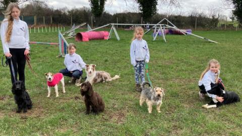 Four children of various ages stand in a dog agility park with a mix of different dogs on leashes. In the background, tunnels, jumps and other obstacles can be seen. 