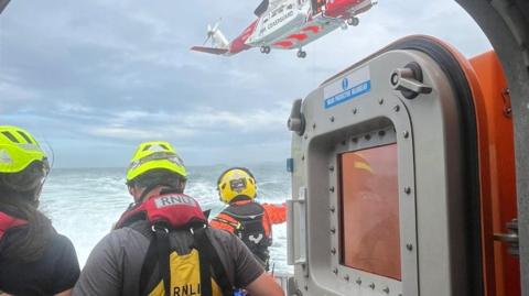 A shot from behind of three people with yellow helmets and life jackets on. They are standing on a boat and looking out the water. A coastguard helicopter is flying above. 