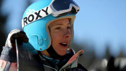 Lesley McKenna is wearing a sky blue helmet with white-rimmed goggles strapped to the top of the helmet. She is holding the top of a snowboard and is looking off to the distance.