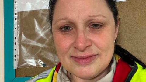 Annabelle Storm has dark hair tied back. She is standing in front of a pinboard and is wearing a high vis jacket over a red top.