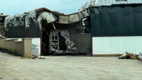 A damaged shed after a fire. A van is burnt out and a roof has partially caved in. It's day time.