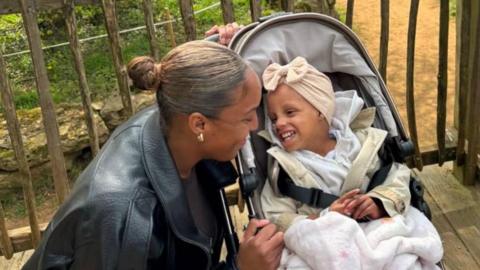 Amarah is smiling at her daughter who is sat in a buggy. Her Daughter, Ariyah is wearing a bowtie head wrap around her head and has blankets over her. She is smiling at her mother. 