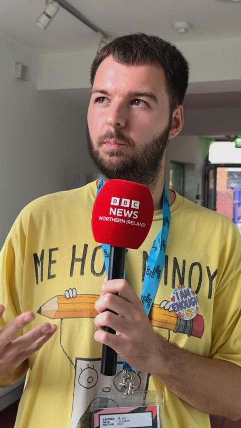 A man with short brown hair and a beard wearing a yellow tshirt - he is holding a microphone with a red cover in it which has BBC NEWS NORTHERN IRELAND on it in white text